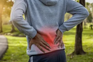 Person experiencing lower back pain while standing outdoors in a park, with a prominent red overlay highlighting the area of discomfort.